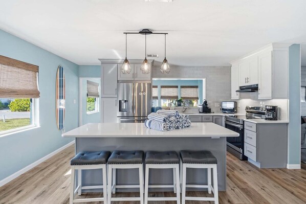 A modern kitchen with a central island, bar stools, stainless steel appliances, and a view to the outside. - Sea Salt & Sunny Days: Pool, Spa, Putting Green by Anna Maria Life Vacation Rentals (Holmes Beach)