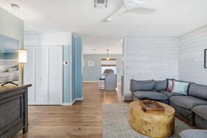 A modern living room with a gray sofa, a wooden coffee table, and a view of a kitchen area. - Sea Salt & Sunny Days: Pool, Spa, Putting Green by Anna Maria Life Vacation Rentals (Holmes Beach)