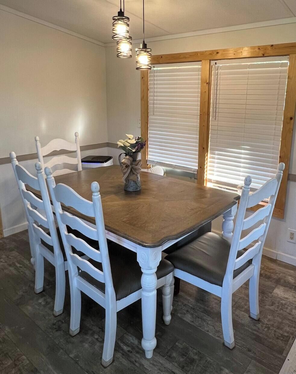 Un coin repas avec une table en bois et des chaises blanches, un vase rempli de fleurs et une fenêtre avec des stores.