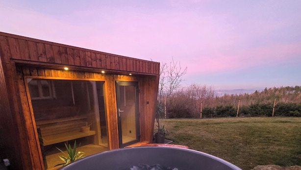 Ein Whirlpool mit einer Holzdielen-Terrasse, ein Holzhaus mit Glasüren und Blick auf die Landschaft.
