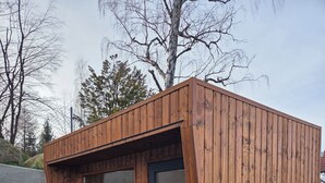 Eine moderne Holzkonstruktion mit großen Glasüren, einer Betonwand und einem Grünbereich mit Topfpflanzen.