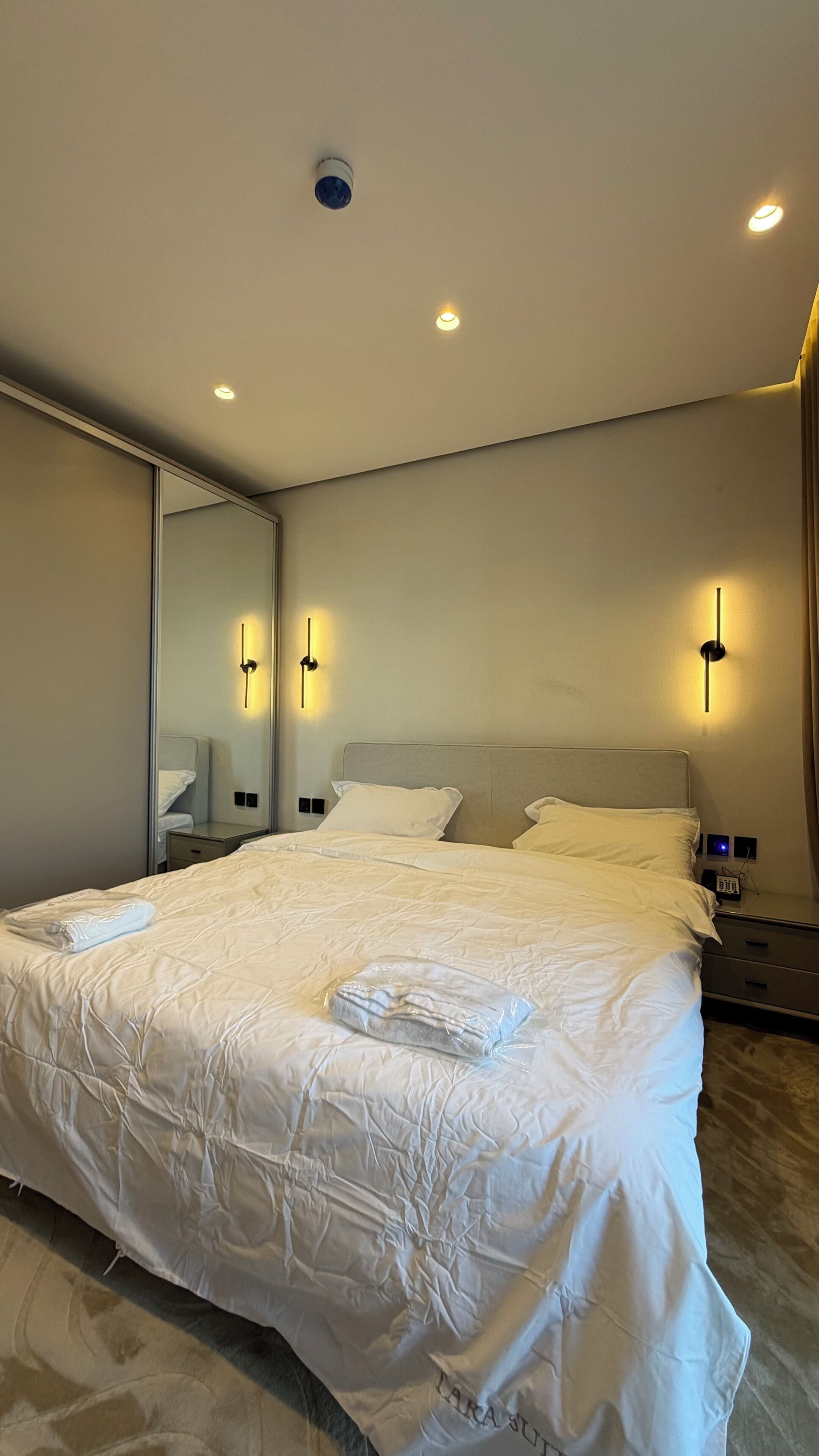 A hotel room with a large bed, bedside tables, a mirror, and wall-mounted lighting.