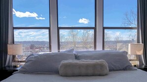A bed with two pillows and a fluffy blanket, a window with a city view, and two bedside lamps. - City View Perch Walkable Apt (Cincinnati)