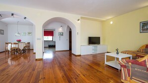 Zona d'estar oberta amb sòl de fusta, taula de dinar amb caires, sofà, televisor i un armari blanc.