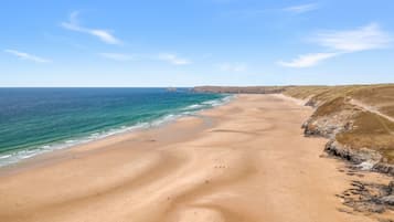 A wide sandy beach with gentle waves, a clear sky, and rocky cliffs in the background.