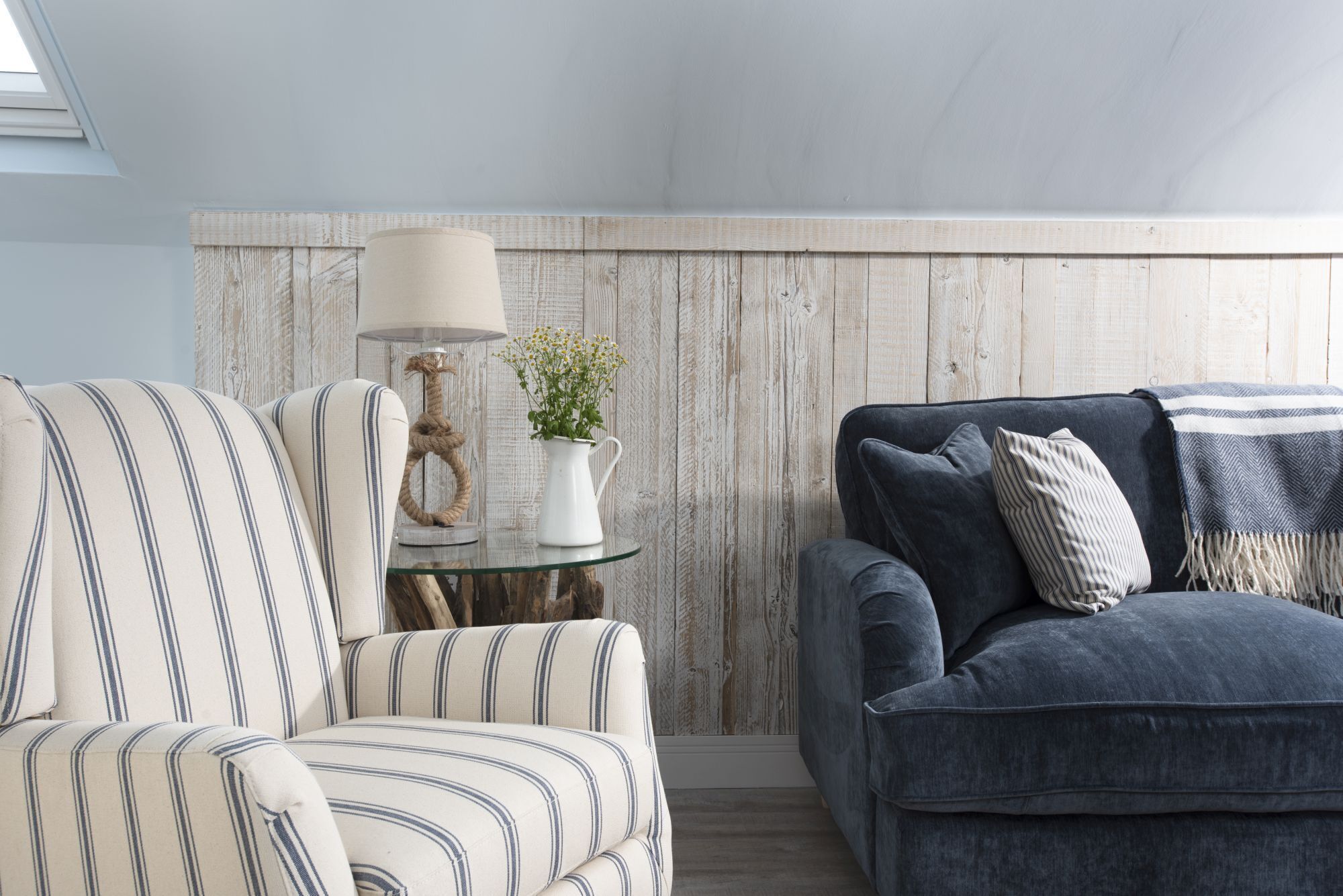 A living room with striped armchairs, a dark sofa, a glass coffee table with a lamp and a plant, and a wooden wall.