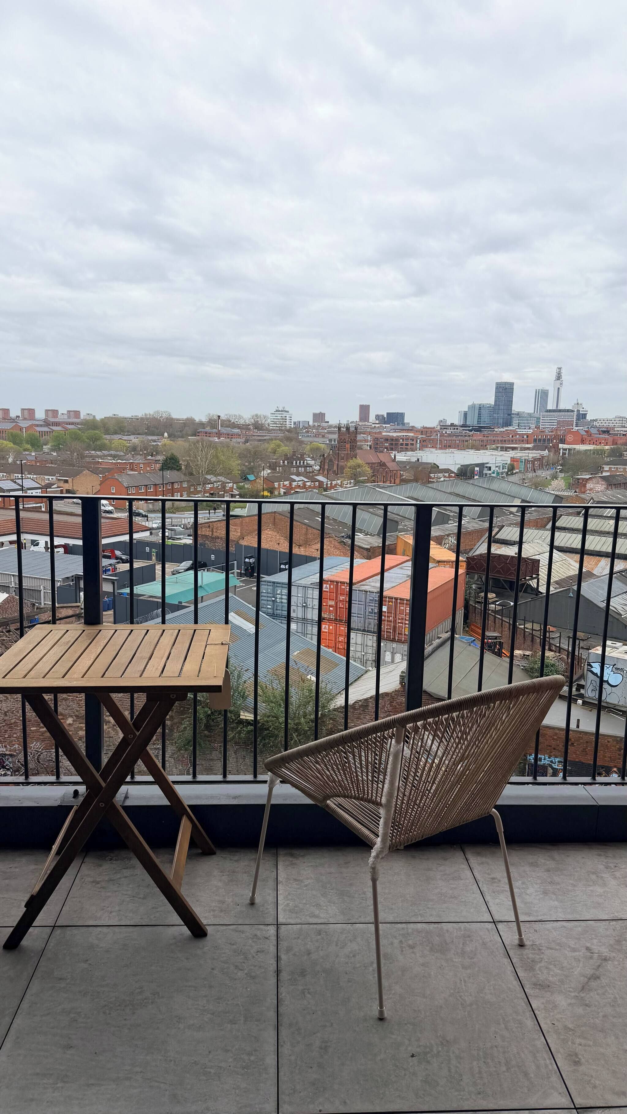 A balcony with a table and chair, overlooking a cityscape with buildings and a clear sky.