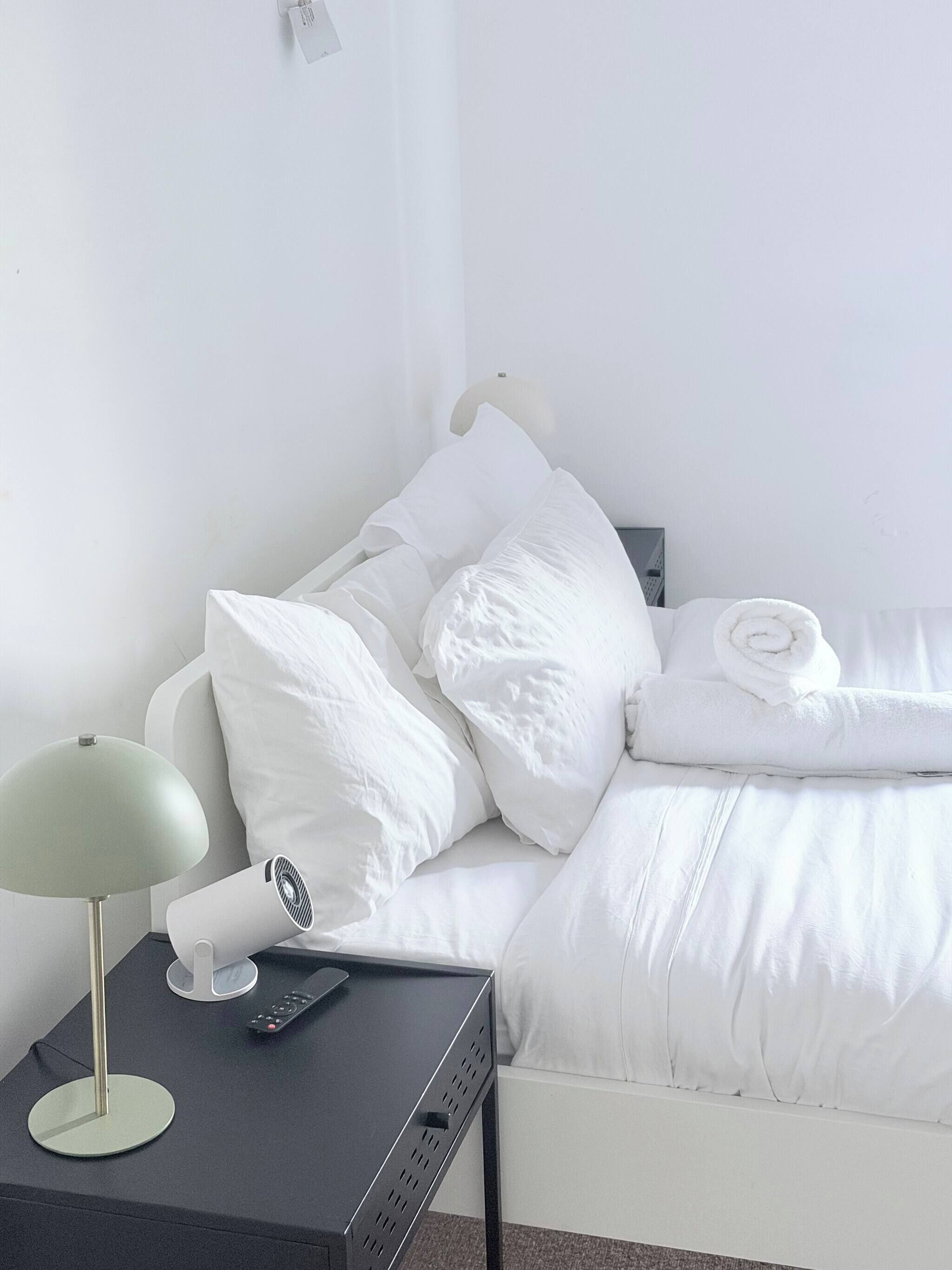 Een net opgemaakt bed met witte lakens, een nachtkastje met een lamp en een ventilator, en een afstandsbediening op tafel.