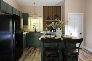 Cocina con armarios verdes, refrigerador negro, isla de madera con dos sillas, lámpara colgante y puerta blanca.