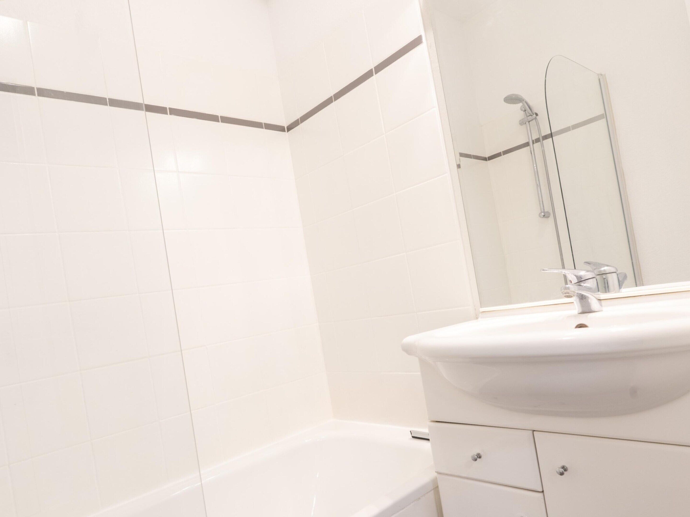 A bathroom with white tiles, a white sink, a mirror, and a shower area.