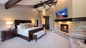 Een slaapkamer met een groot bed, een nachtkastje, een bankje, een open haard, een plafondventilator en een aan de muur bevestigde televisie.