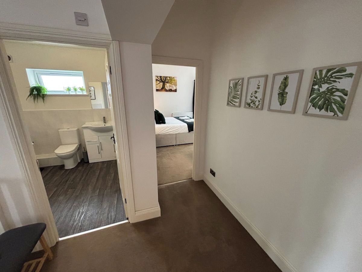 Un couloir mène à une chambre à coucher où se trouve un lit, ainsi qu’à une salle de bain équipée de toilettes et d’un lavabo. Des reproductions botaniques encadrées ornent le mur.