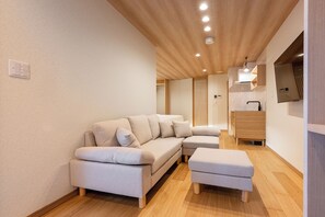 Ein modernes Wohnzimmer mit Sofa, Hocker, Holzboden, deckenebener Beleuchtung und einem integrierten Fernseher.