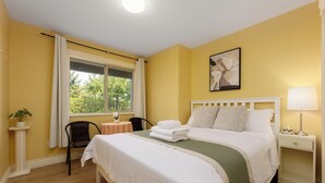 A hotel room with a bed, a bedside table with a lamp, a window with curtains, a chair, and a small table with a plant.