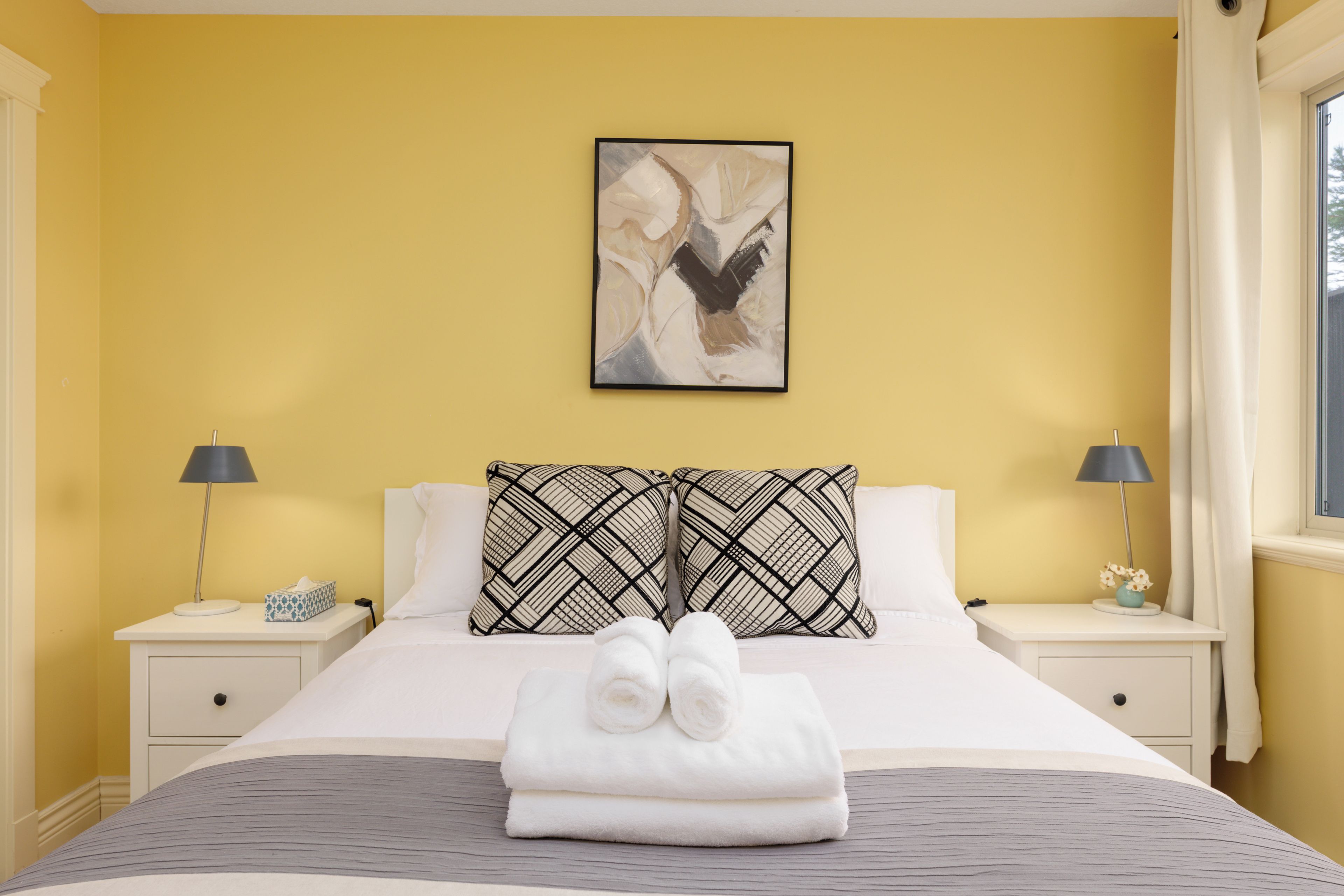 Una camera d'albergo con un letto, due comodini, un quadro e una finestra con tende.