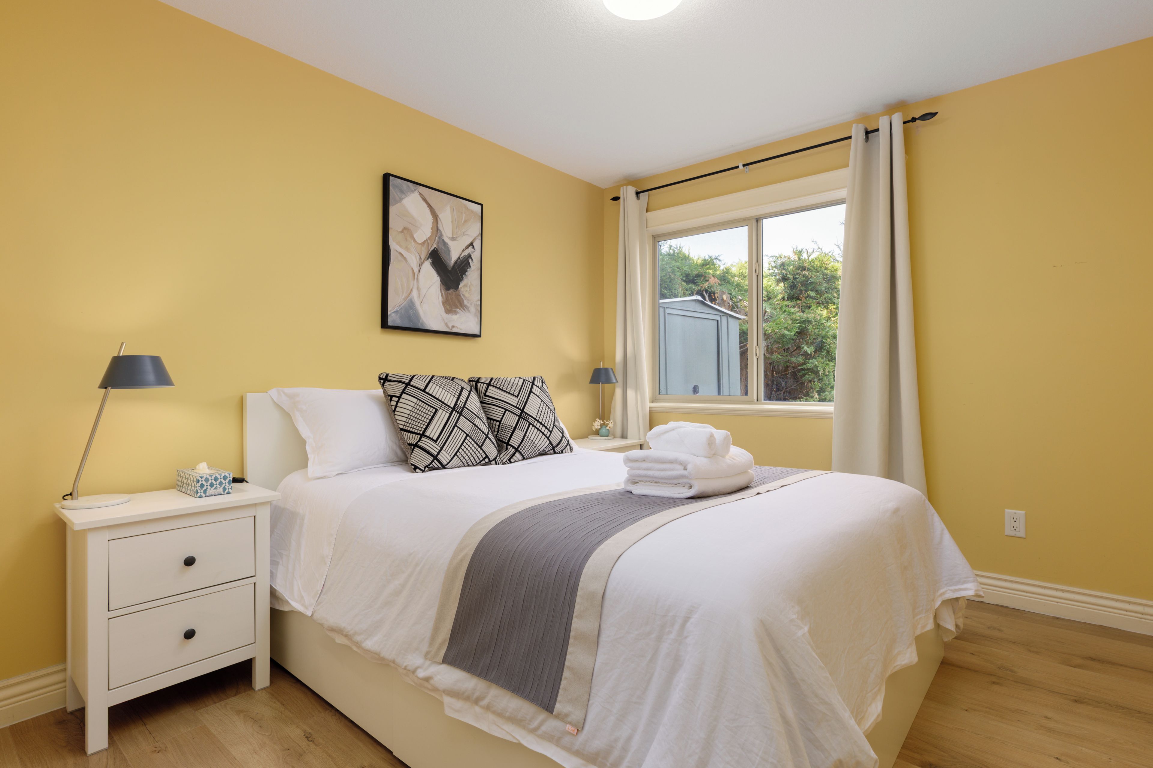 Une chambre à coucher avec un lit, des tables de chevet, une lampe, un tableau et une fenêtre avec des rideaux.