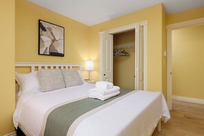 Un lit bien fait, avec du linge de lit blanc et un plaid vert, une lampe de chevet et un tableau accroché au mur.