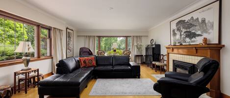 A spacious living room with a black leather sofa, a fireplace, and a large window offering a view of greenery.
