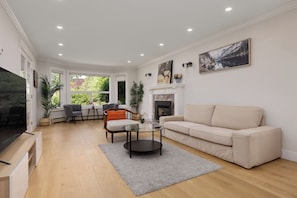 A modern living room with a beige sofa, a glass coffee table, and a fireplace. There is a dining area with a table and chairs, and a large window providing natural light. - Spacious 6BR Home Near DT and 15 mins to YVR (Vancouver)