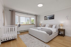 Ein Schlafzimmer mit einem Bett, einem Babybett, einem Fenster mit Jalousien, einer Nachttischlampe und einem Wandbild.