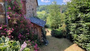 Ein rustikales Steinhaus mit überdachter Veranda, umgeben von üppigem Grün und blühenden Pflanzen.