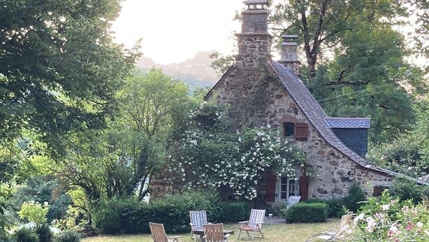 Ein Steinhäuschen mit Schornstein, umgeben von üppiger Vegetation und einer grünen Wiese mit vier Holzstühlen.