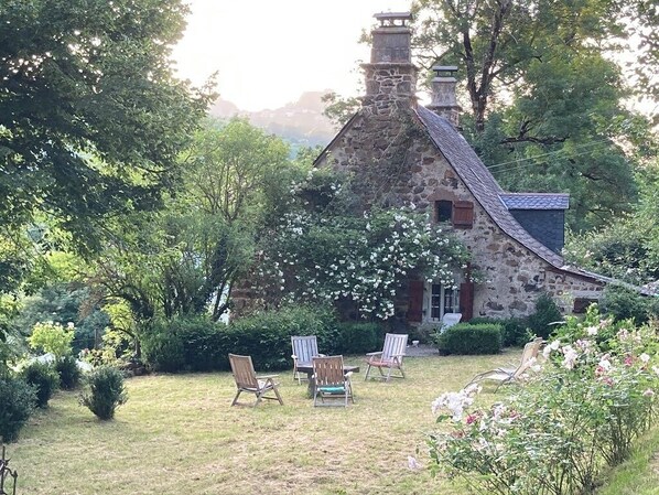 A stone house with a chimney, surrounded by lush greenery and a grassy lawn with four wooden chairs. - Volcanic Retreat - character house in the heart of Cantal, nature & silence. (Saint-Hippolyte)