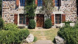 Ein Steinhäuschen mit mittlerem Eingang, hölzernen Fensterläden und einem kleinen Garten mit Sträuchern und einem Weg.