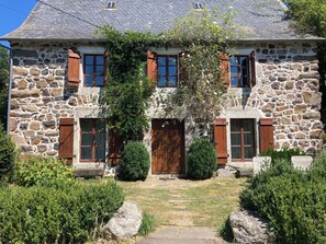 A stone house with a central entrance, wooden shutters, and a small garden with shrubs and a pathway. - Volcanic Retreat - character house in the heart of Cantal, nature & silence. (Saint-Hippolyte)