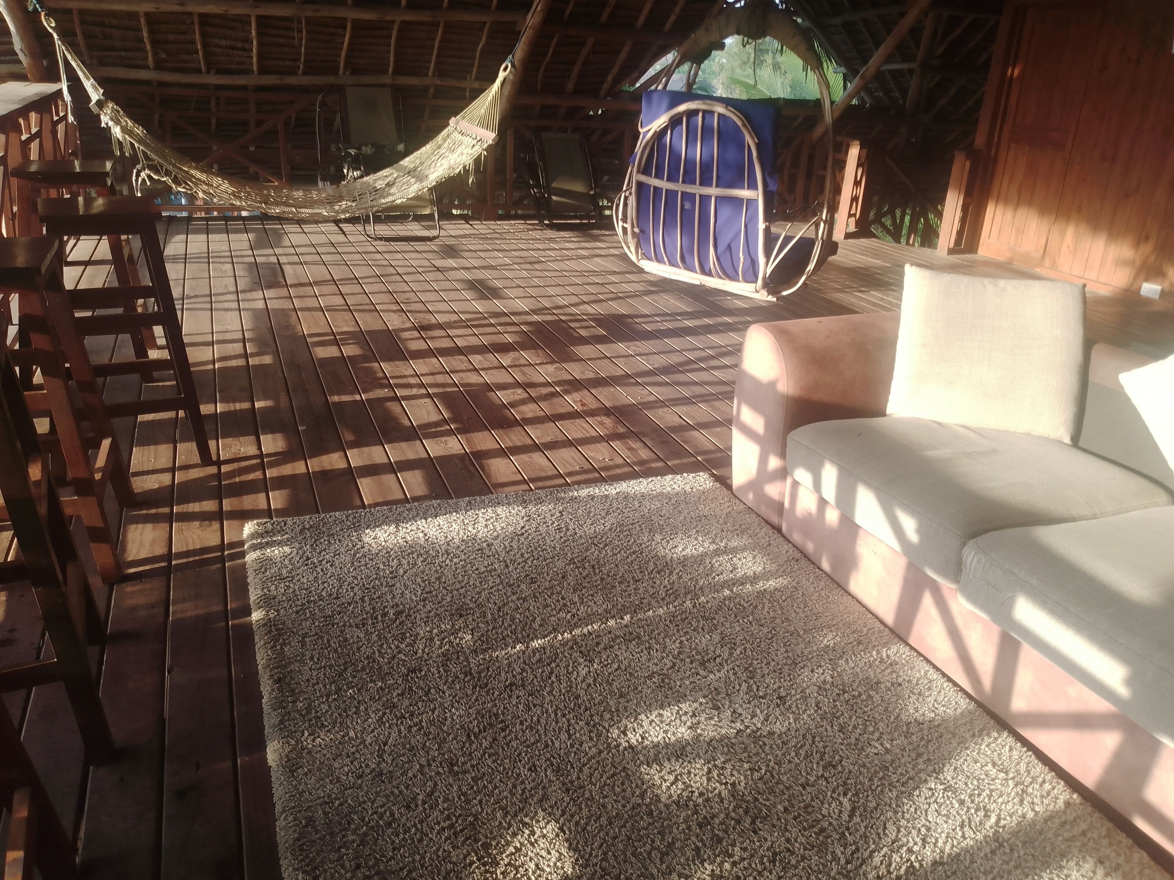 Una hamaca, una silla acolchada y un sofá en una terraza de madera con techo de paja.