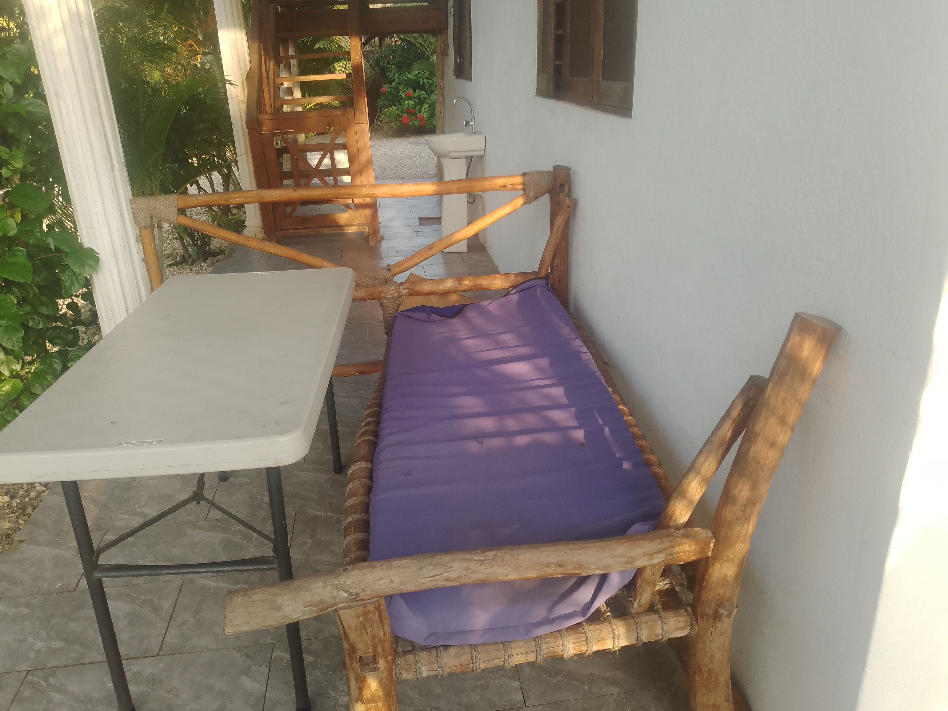 Un banco de madera con un cojín morado, una mesa blanca y una ventana con vista a la vegetación.