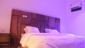 A bedroom with a tufted headboard, a bed with white bedding, a nightstand, and an air conditioning unit on the wall.