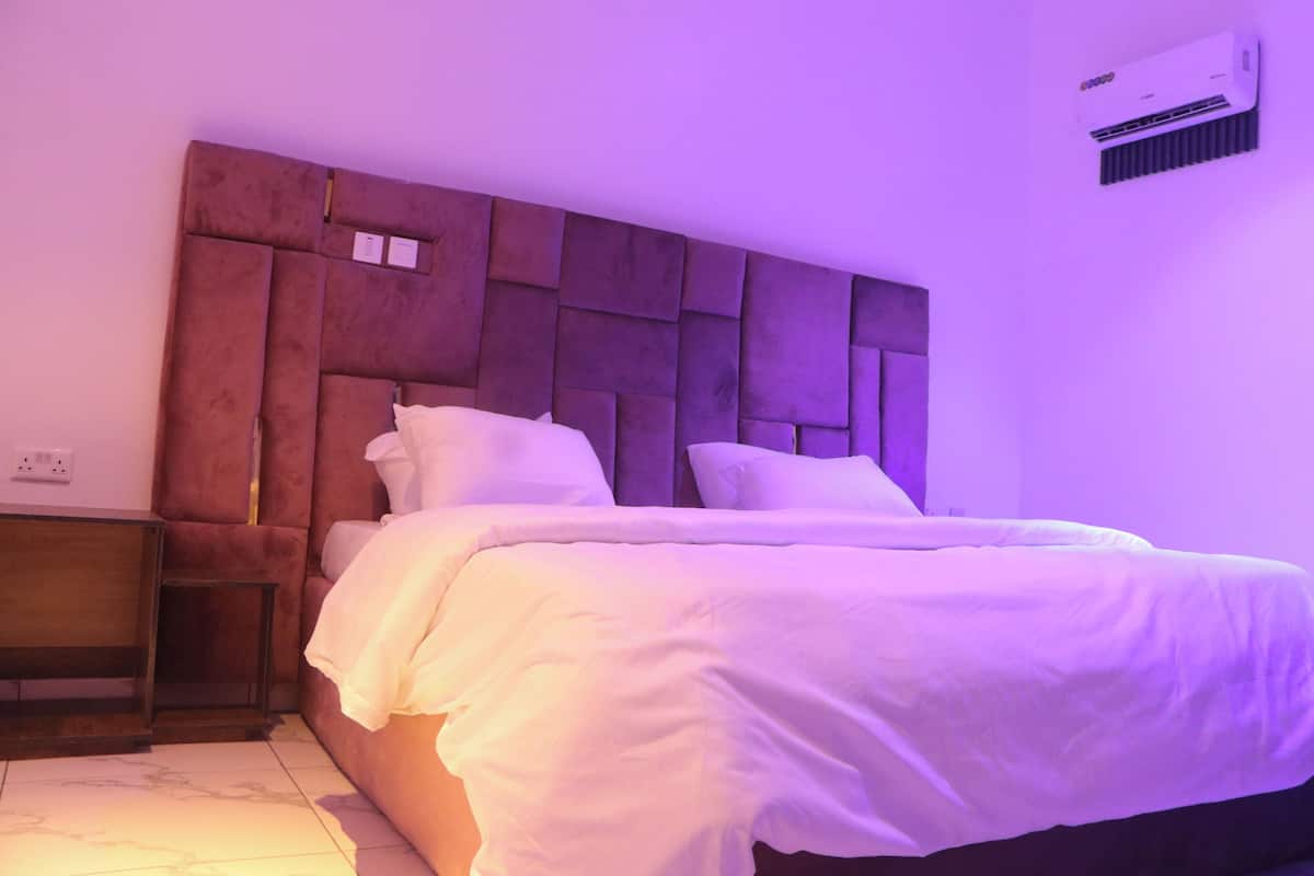 A bedroom with a tufted headboard, a bed with white bedding, a nightstand, and an air conditioning unit on the wall.