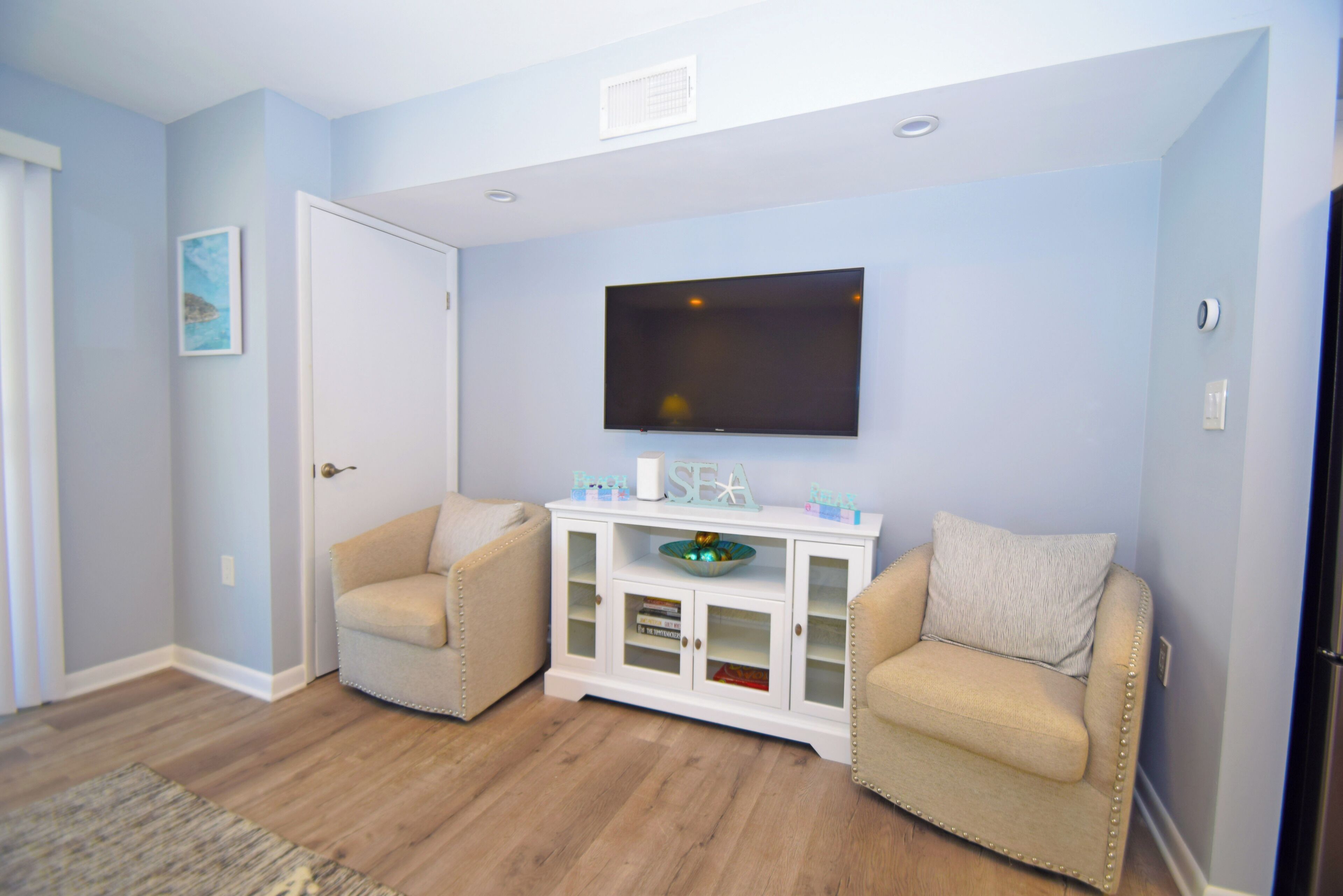 Un salon avec deux fauteuils beiges, un meuble de rangement blanc intégrant un téléviseur à écran plat, et une porte.