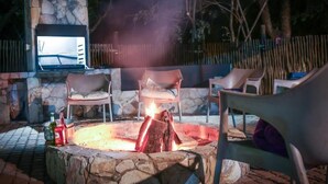 Une cheminée en pierre avec un feu, entourée de chaises d’extérieur et d’un écran de télévision.