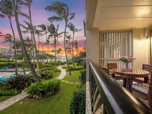 A balcony with a view of a landscaped garden, a pool area, and a sunset over the ocean.