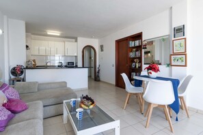 A modern living room with a sofa, coffee table, dining table with chairs, a kitchen in the background, and a bookshelf with books and decorations. - Paloma Beach 15 - One Bed Penthouse (Los Cristianos)