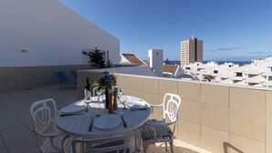 Eine Dachterrasse mit einem runden Esstisch, weißen Stühlen und Blick auf das Meer und die Stadt.