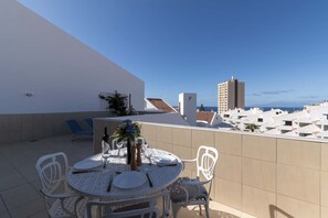 A rooftop terrace with a round table set for dining, white chairs, and a view of the sea and buildings. - Paloma Beach 15 - One Bed Penthouse (Los Cristianos)