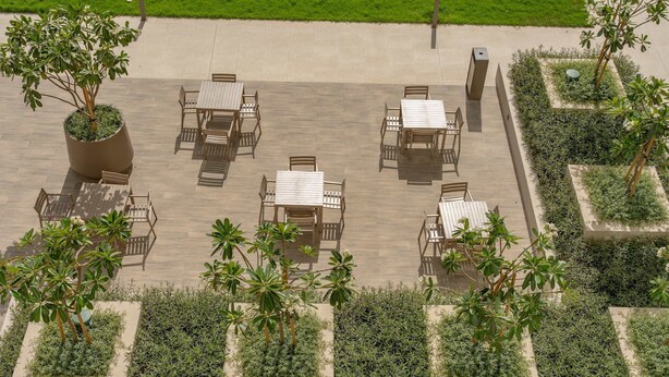Un patio exterior con varias mesas y sillas, rodeado de follaje y un camino pavimentado.