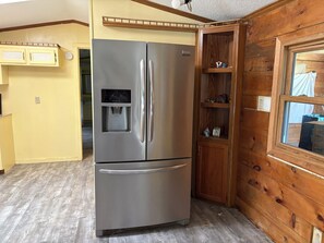 A stainless steel refrigerator with a water dispenser, a wooden cabinet with shelves, and a window with a view. - Rustic Lake country 3-bed 2 bath home on beautiful road 2.5 mile to Otisco Lake. (Marietta)