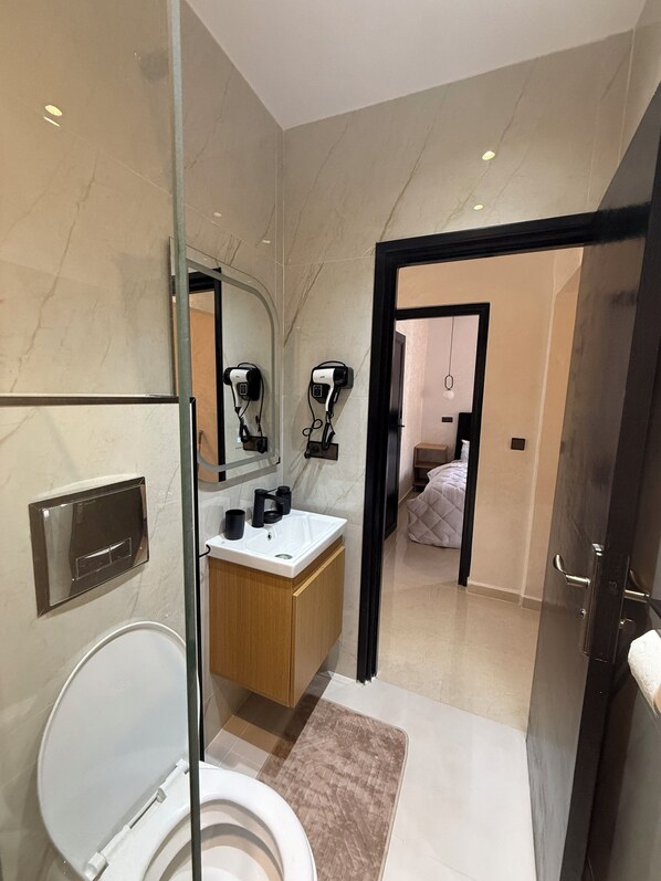 Une salle de bain moderne dotée d’une douche à parois de verre, de toilettes blanches, d’un meuble-lavabo en bois avec un miroir et d’un sèche-cheveux fixé au mur.