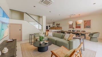 A modern living room with a green sofa, a coffee table, and a wicker chair. There is a staircase leading to an upper level.