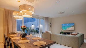 Una sala de estar moderna con comedor, un conjunto de sofás y un televisor mostrando una escena de playa.