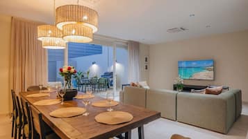 A modern living room with a dining area, a sofa set, and a TV displaying a beach scene.