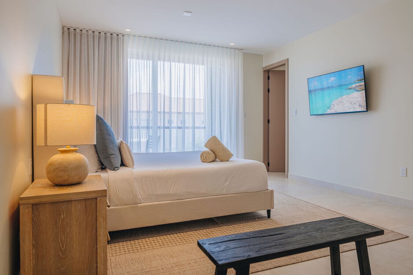A hotel room with a bed, a bedside lamp, a small table, a painting on the wall, and a view through the window.