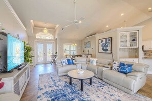 A spacious living room with a large sectional sofa, a flat-screen TV, and a dining area in the background. - Game room - sleeps 8 with large pool! (Cape Coral)