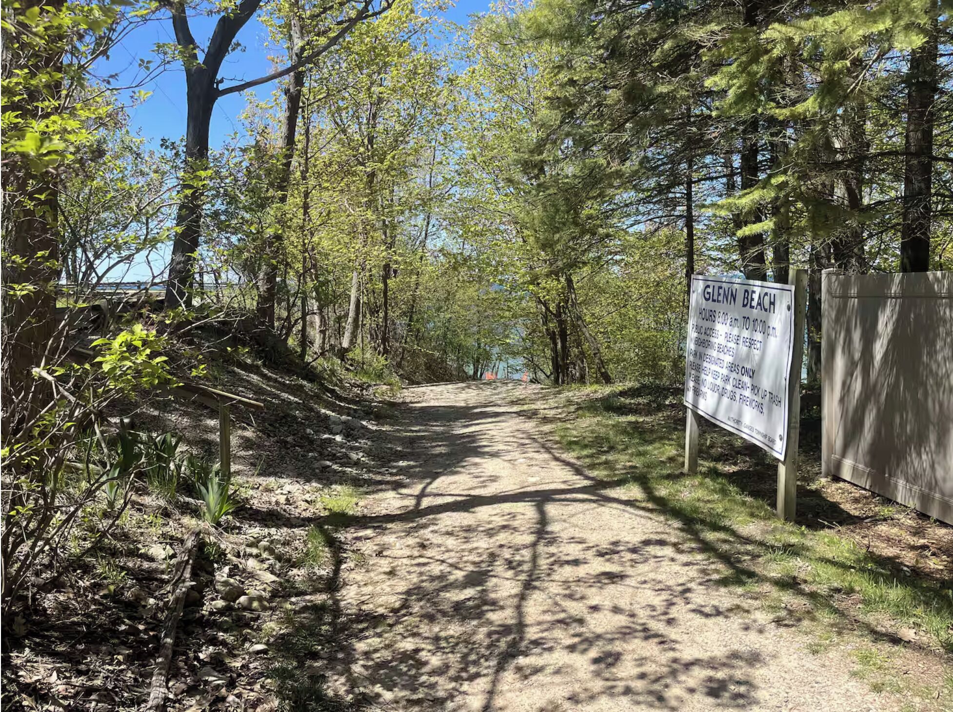 Un sentier de terre mène à une plage, et un panneau indique les heures d’ouverture de la plage et les règles d’accès au public.