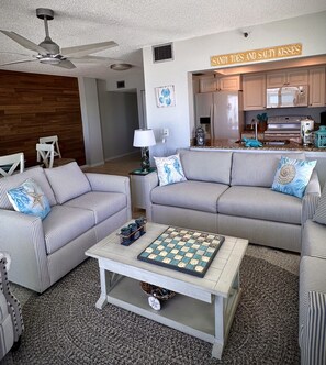 Ein Wohnzimmer mit gestreiften Sofas, einem Couchtisch mit Schachbrett, einem Deckenventilator und einer im Hintergrund sichtbaren Küche.