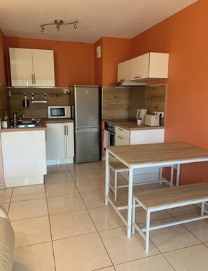 Una cocina compacta con armarios blancos, un refrigerador de acero inoxidable, un microondas y una mesa de madera.